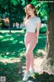 A woman in a white top and pink leggings leaning against a tree.